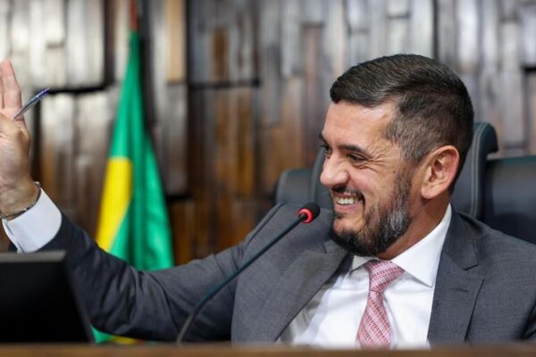 Rodrigo Bacellar em sessão, segurando caneta e sorrindo, com bandeira ao fundo.