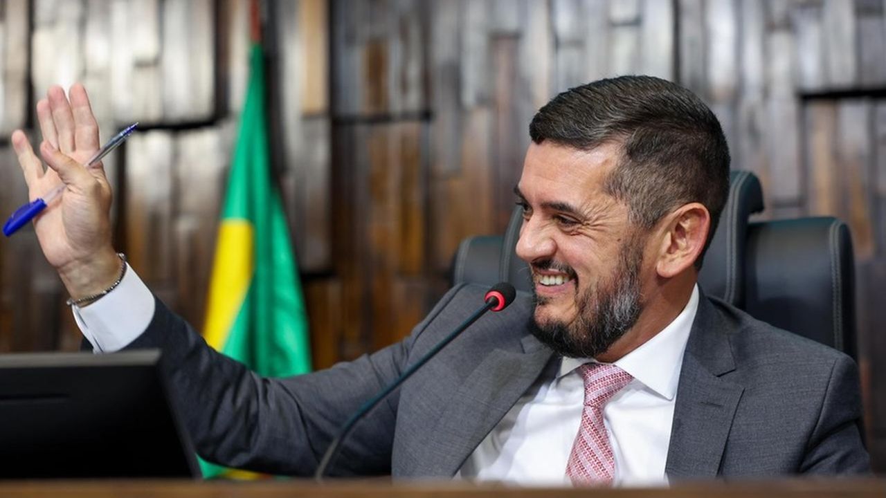 Rodrigo Bacellar em sessão, segurando caneta e sorrindo, com bandeira ao fundo.