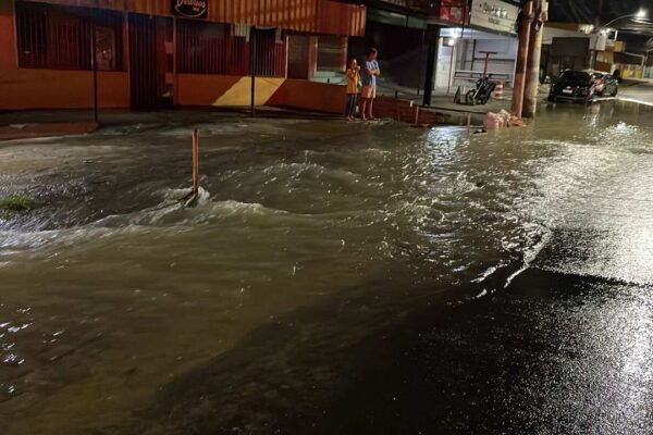 Rua alagada com água corrente, pessoas observando e comércios ao fundo.
