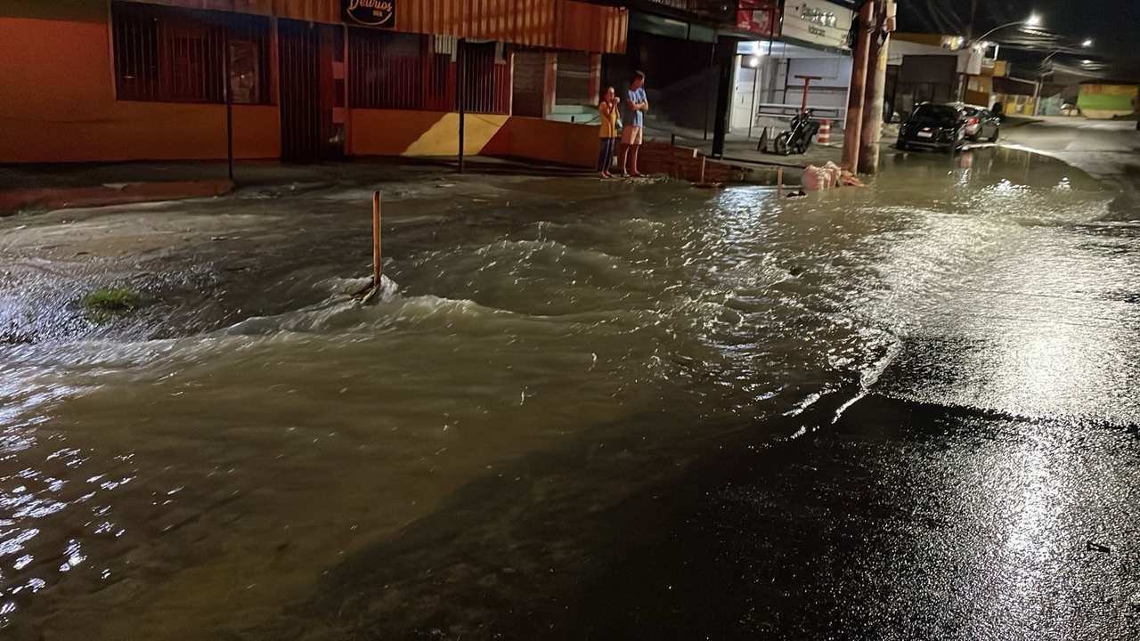 Rua alagada com água corrente, pessoas observando e comércios ao fundo.