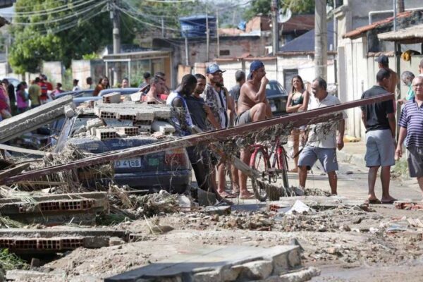 Rua com destroços após fortes chuvas na Baixada Fluminense. Pessoas observam os danos.