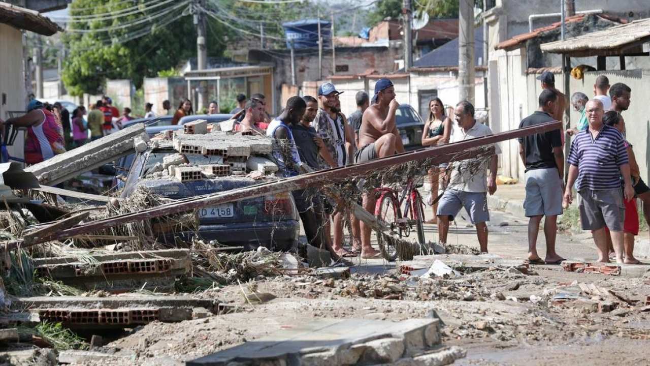 Rua com destroços após fortes chuvas na Baixada Fluminense. Pessoas observam os danos.