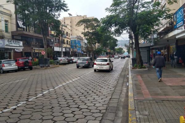 Rua de Nova Friburgo com carros estacionados e prédios comerciais, um homem anda na calçada e um hidrante vermelho em primeiro plano.