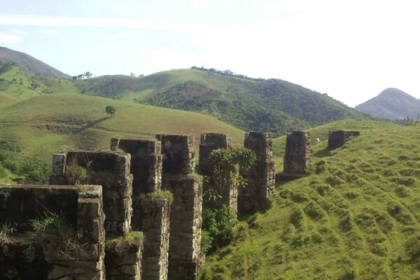 Ruínas de ponte ferroviária em meio a colinas verdes, com vegetação e céu claro.