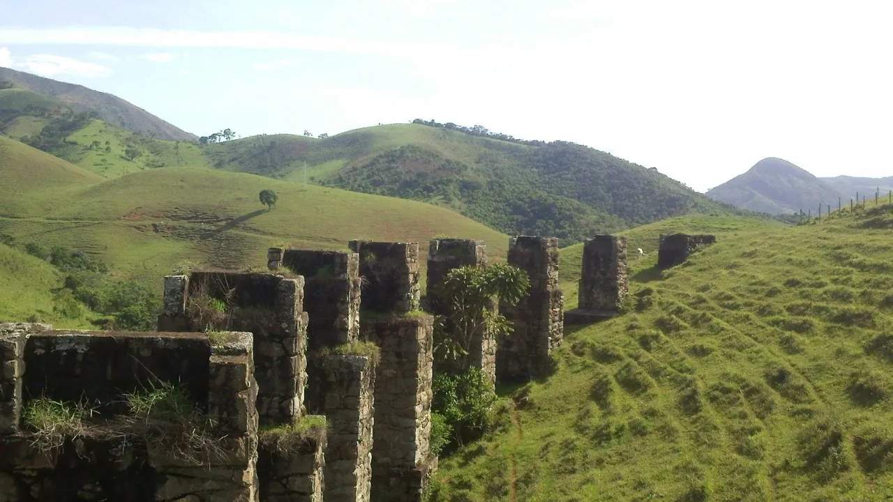 Ruínas de ponte ferroviária em meio a colinas verdes, com vegetação e céu claro.