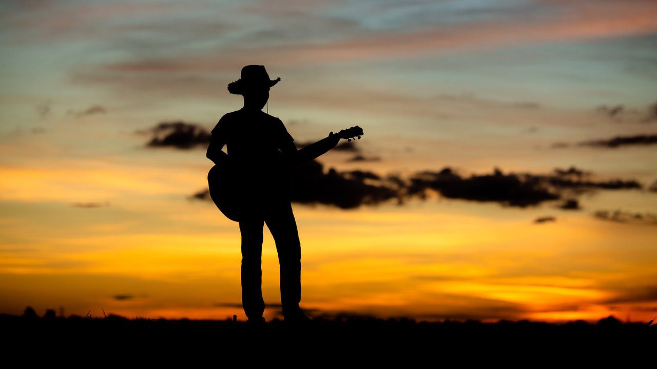 Silhueta de homem tocando violão no pôr do sol, representando artistas do sertanejo.