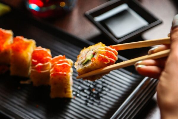 Sushi, close-up de um rolinho sendo pego com hashi, acompanhado por outros rolinhos e molheira.