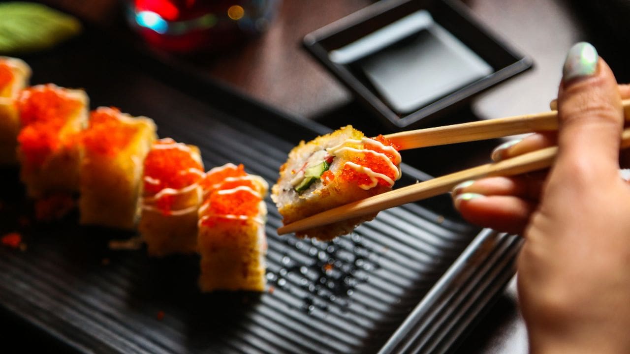 Sushi, close-up de um rolinho sendo pego com hashi, acompanhado por outros rolinhos e molheira.