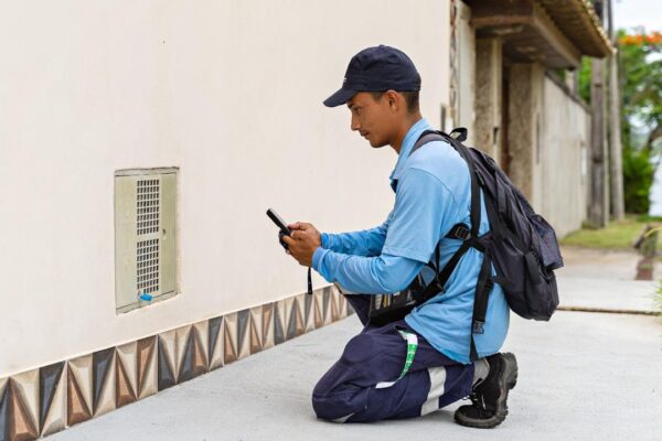 Técnico da Águas de Nova Friburgo, uniformizado, analisa hidrômetro com celular, em frente a uma residência.