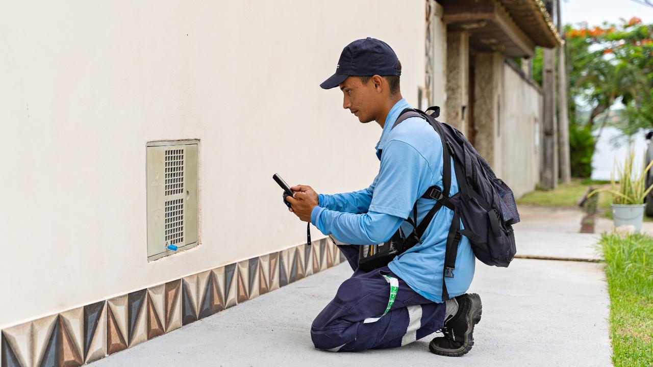 Técnico da Águas de Nova Friburgo, uniformizado, analisa hidrômetro com celular, em frente a uma residência.