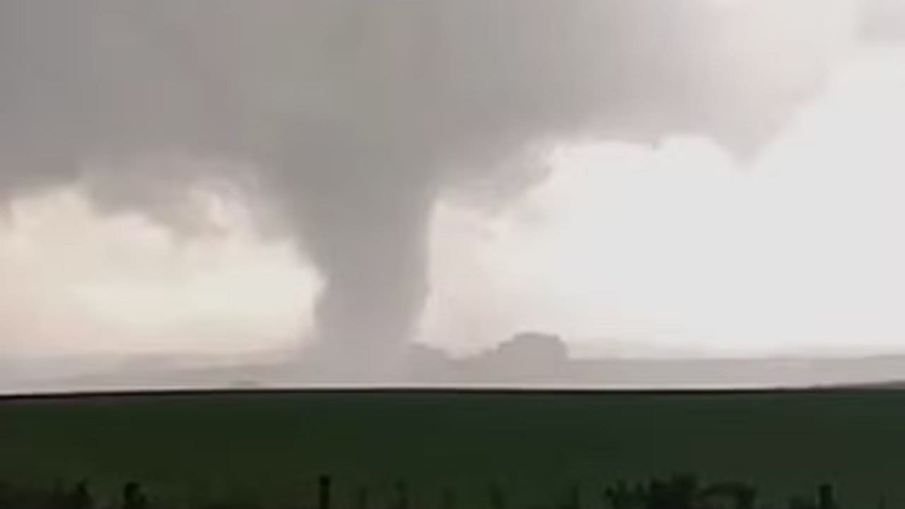 Tornado passando por uma área rural no Rio Grande do Sul, com céu nublado e uma grande coluna de ar girando em direção ao chão.