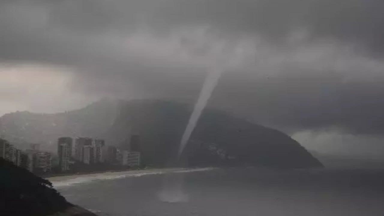 Tornado sobre a praia no Rio de Janeiro durante tempestade, com prédios e morro ao fundo.