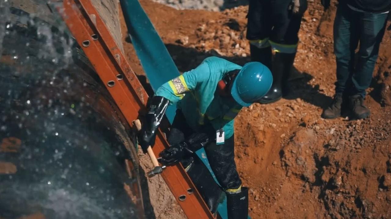 Trabalhador em manutenção de tubulação de água, usando martelo para reparo, com água jorrando do cano.