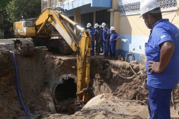 Trabalhadores em uma obra de manutenção de rede de água, com escavadeira e canos expostos.