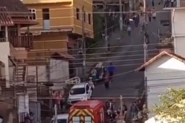 Uma cena caótica com vários carros, um caminhão de bombeiros e pessoas reunidas em uma rua de paralelepípedos.