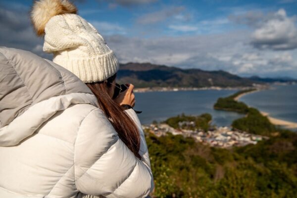 Uma pessoa com um casaco branco e chapéu de lã observa a paisagem montanhosa e o mar ao fundo.