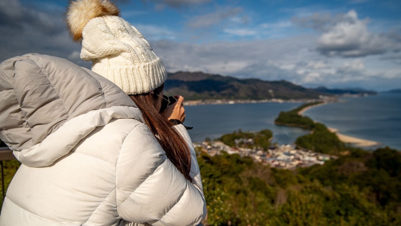 Uma pessoa com um casaco branco e chapéu de lã observa a paisagem montanhosa e o mar ao fundo.