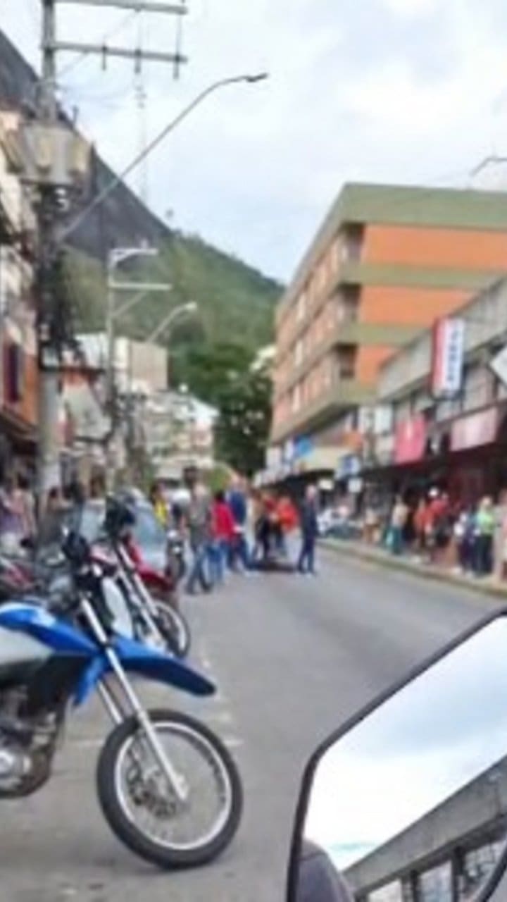 Uma rua com várias motocicletas estacionadas, uma mulher caída no chão e pessoas ao redor, em Nova Friburgo, RJ.