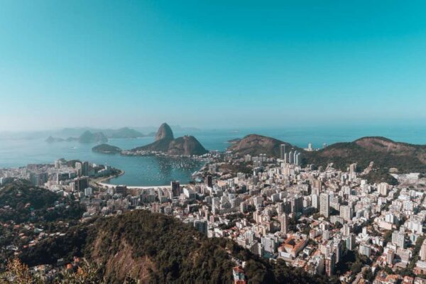 Vista aérea da cidade do Rio de Janeiro com o mar ao fundo, sob um céu azul claro.
