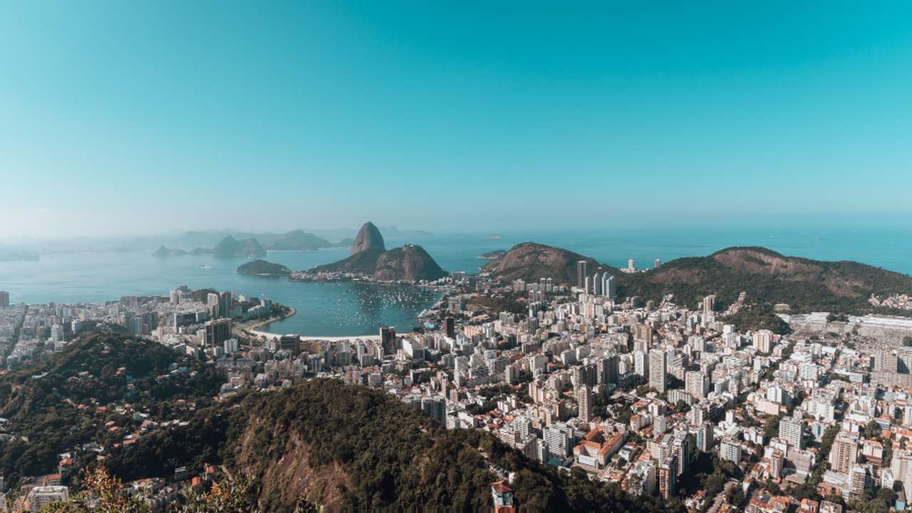 Vista aérea da cidade do Rio de Janeiro com o mar ao fundo, sob um céu azul claro.