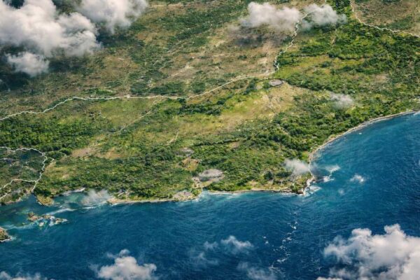 Vista aérea da costa com vegetação densa e mar azul, sob céu parcialmente nublado.