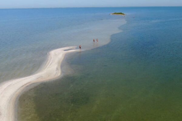 Vista aérea da Ilha do Boi, em São Pedro da Aldeia, com pessoas caminhando na faixa de areia no mar.