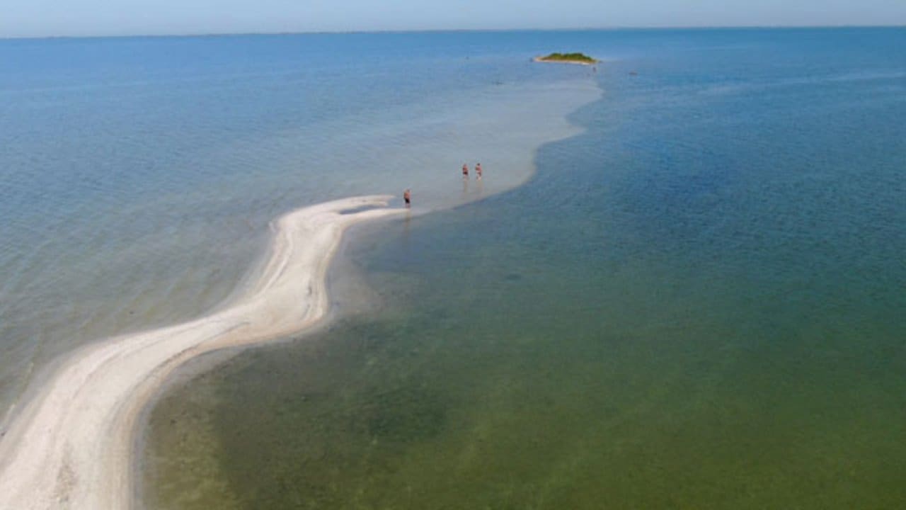 Vista aérea da Ilha do Boi, em São Pedro da Aldeia, com pessoas caminhando na faixa de areia no mar.