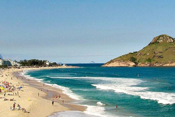 Vista aérea da Praia da Macumba, no Rio de Janeiro, com areia branca, mar azul e um morro verde ao fundo.