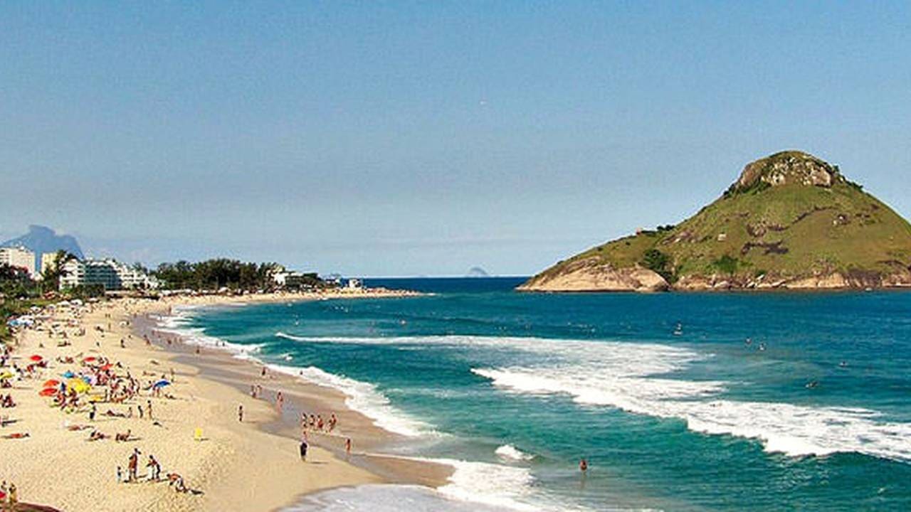 Vista aérea da Praia da Macumba, no Rio de Janeiro, com areia branca, mar azul e um morro verde ao fundo.