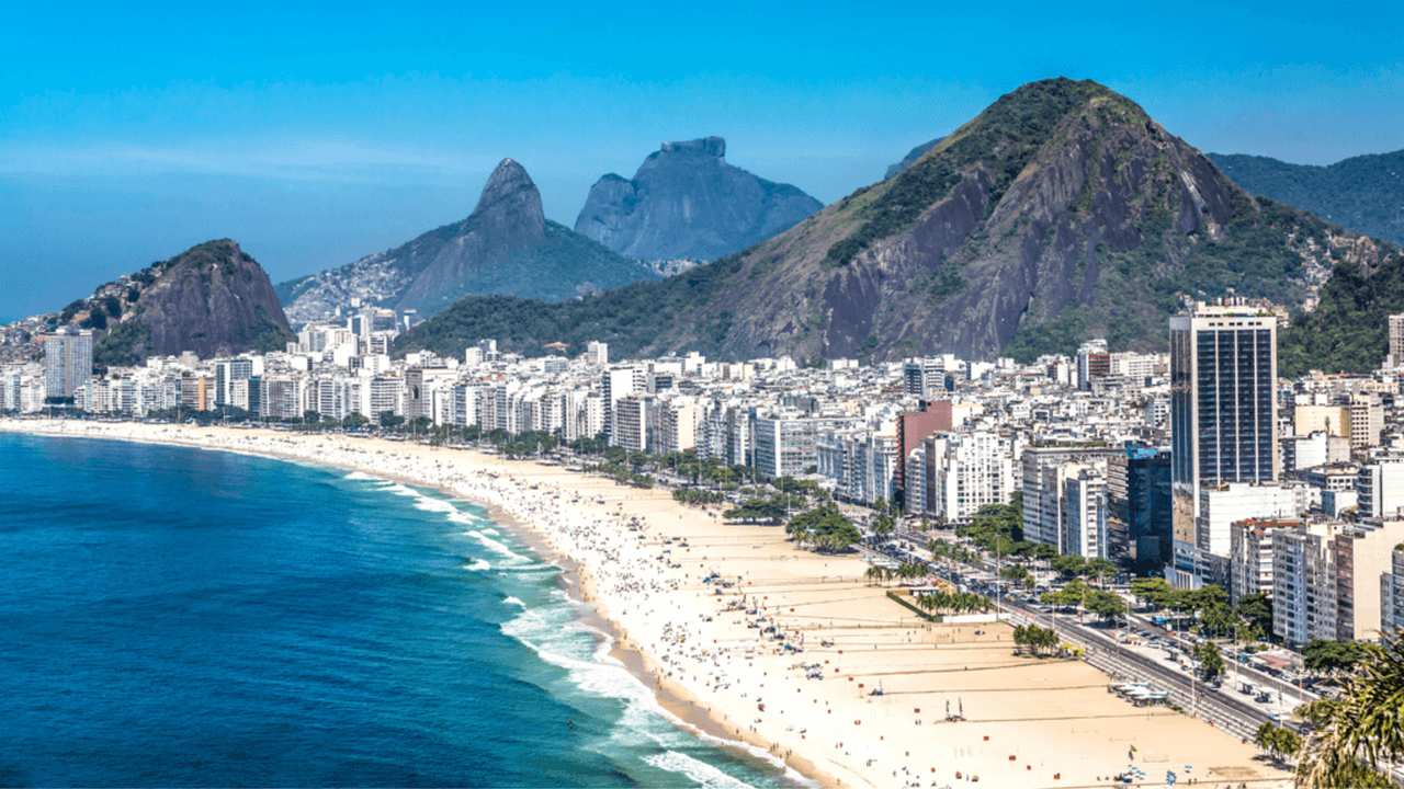 Vista aérea da Praia de Copacabana no Rio de Janeiro, com prédios e montanhas ao fundo.
