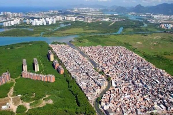 Vista aérea de favela Rio das Pedras em Muzema, Rio de Janeiro, com prédios ao lado e natureza ao fundo.