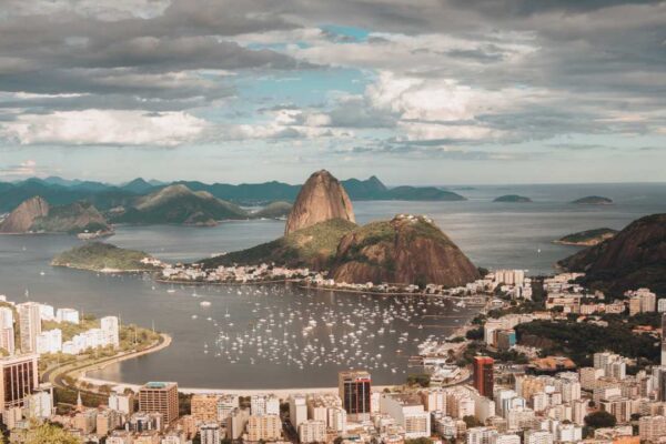 Vista aérea de uma cidade costeira com edifícios, montanhas e uma baía com barcos, sob um céu nublado.