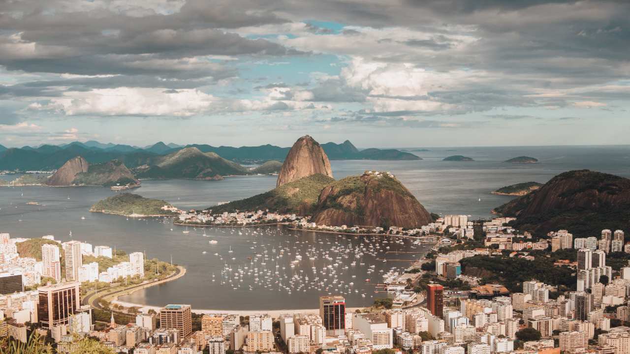 Vista aérea de uma cidade costeira com edifícios, montanhas e uma baía com barcos, sob um céu nublado.
