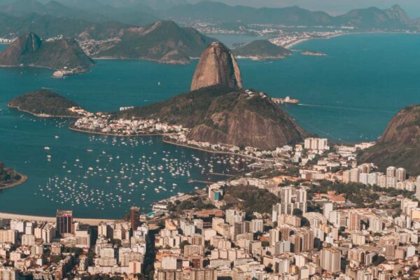 Vista aérea de uma cidade costeira com prédios e montanhas, com mar azul e muitos barcos à vela, sob céu claro.