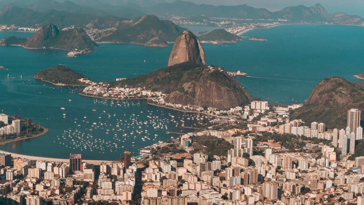 Vista aérea de uma cidade costeira com prédios e montanhas, com mar azul e muitos barcos à vela, sob céu claro.