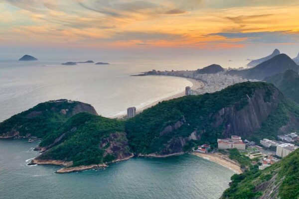 Vista aérea de uma cidade litorânea, com montanhas e oceanos ao fundo. O céu está com nuvens alaranjadas, indicando um pôr do sol.