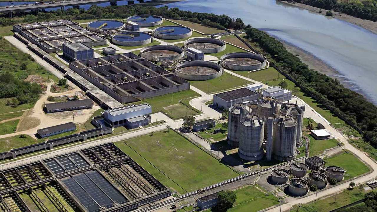 Vista aérea de uma estação de tratamento de água com tanques circulares, filtros retangulares e silos altos, indicando uma grande estrutura de tratamento de água.