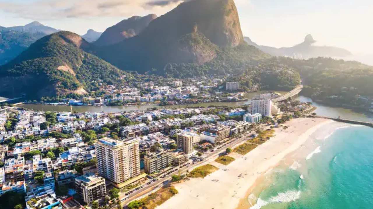 Vista aérea de uma praia no Rio de Janeiro, com edifícios, montanhas e mar azul.