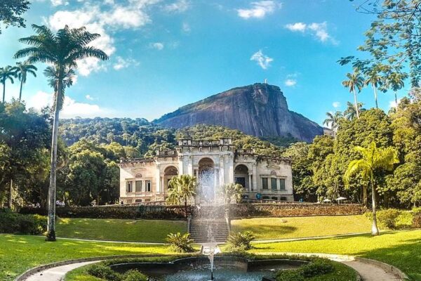 Vista do Parque Lage, no Rio de Janeiro, com o palacete histórico e uma fonte em frente, com a Pedra da Gávea ao fundo.