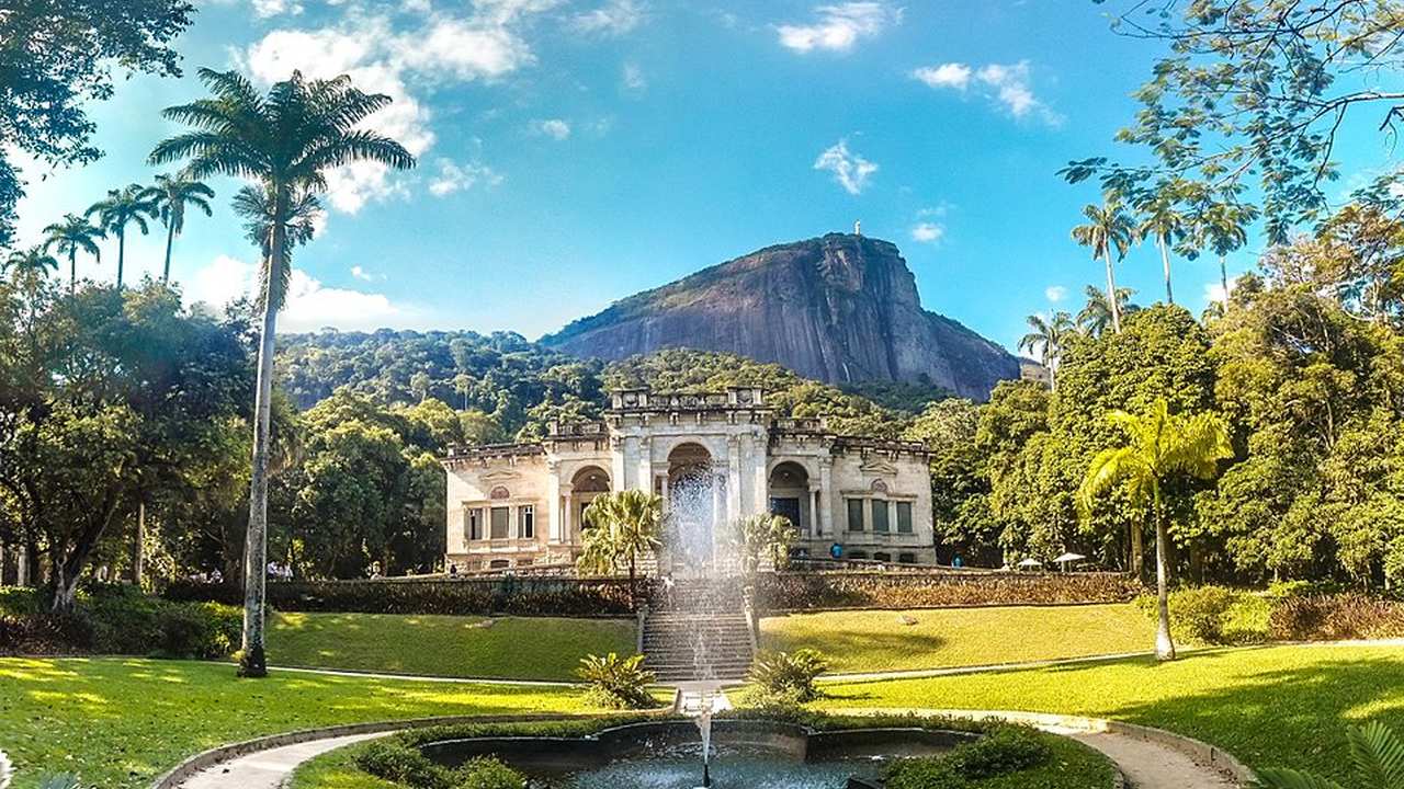 Vista do Parque Lage, no Rio de Janeiro, com o palacete histórico e uma fonte em frente, com a Pedra da Gávea ao fundo.