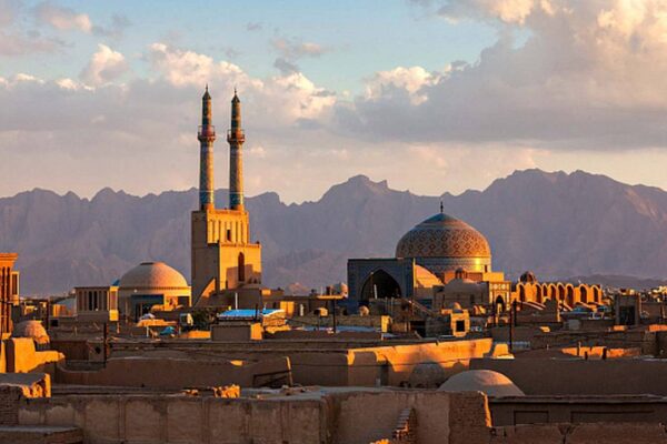 Vista panorâmica de Yazd, Irã, com mesquitas, torres e montanhas ao fundo, sob céu azul.
