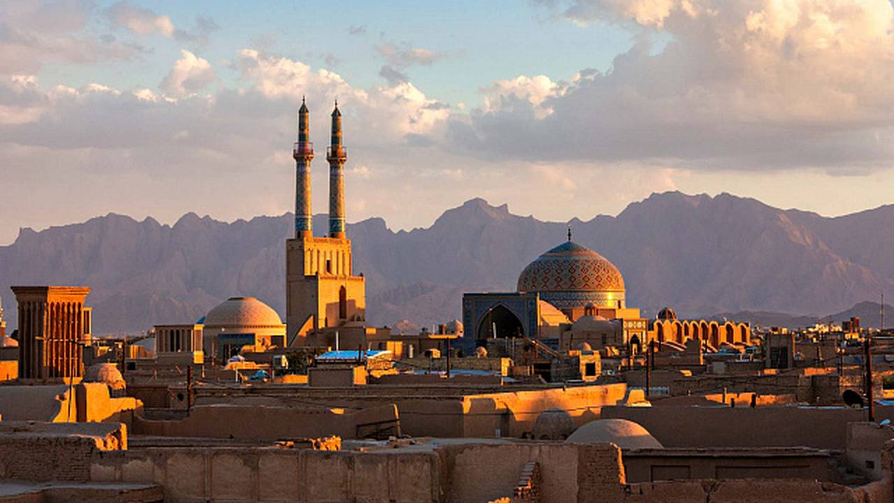 Vista panorâmica de Yazd, Irã, com mesquitas, torres e montanhas ao fundo, sob céu azul.