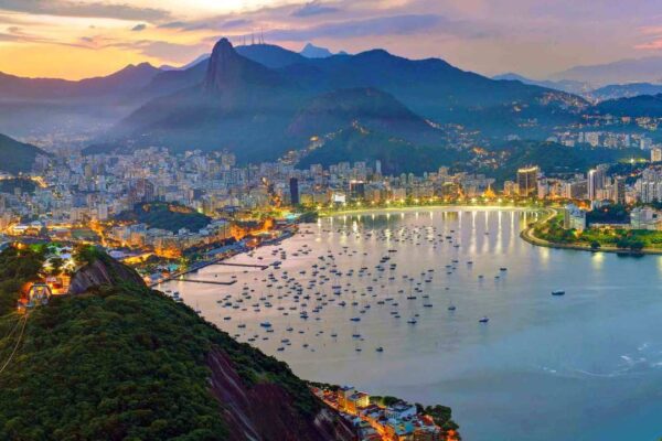 Vista panorâmica do Rio de Janeiro ao entardecer, com montanhas, cidade iluminada e mar.