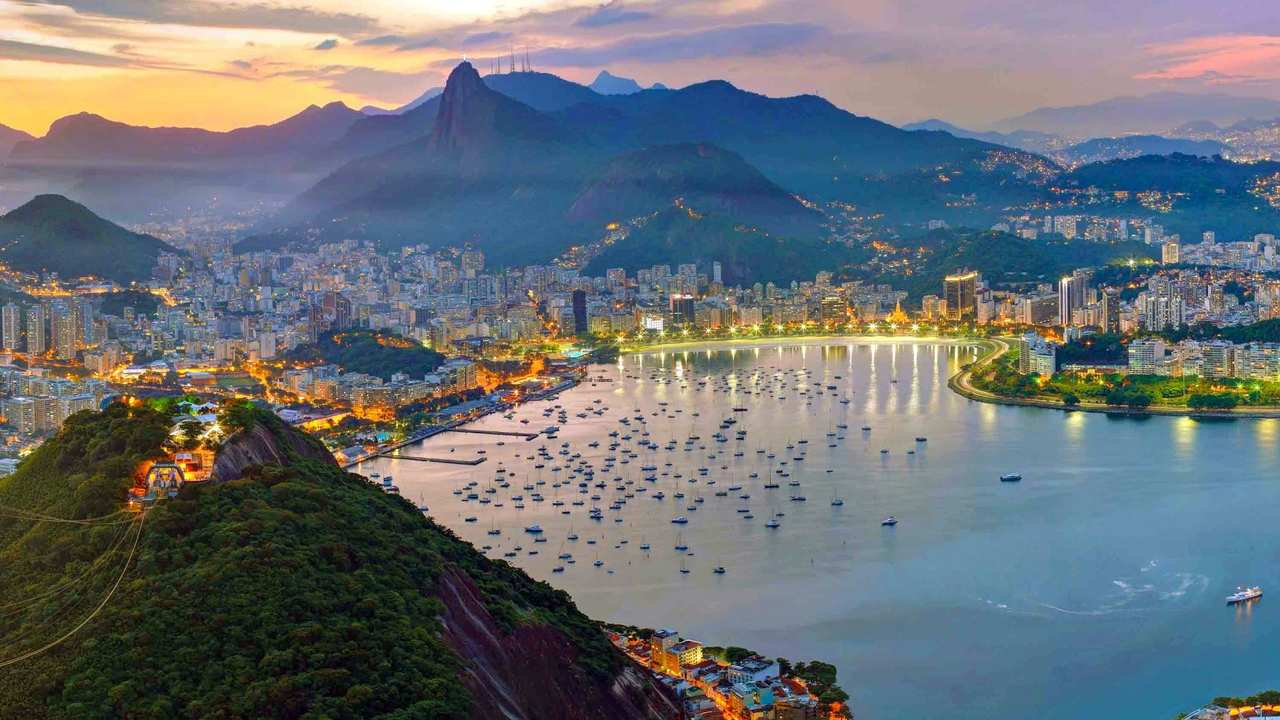 Vista panorâmica do Rio de Janeiro ao entardecer, com montanhas, cidade iluminada e mar.