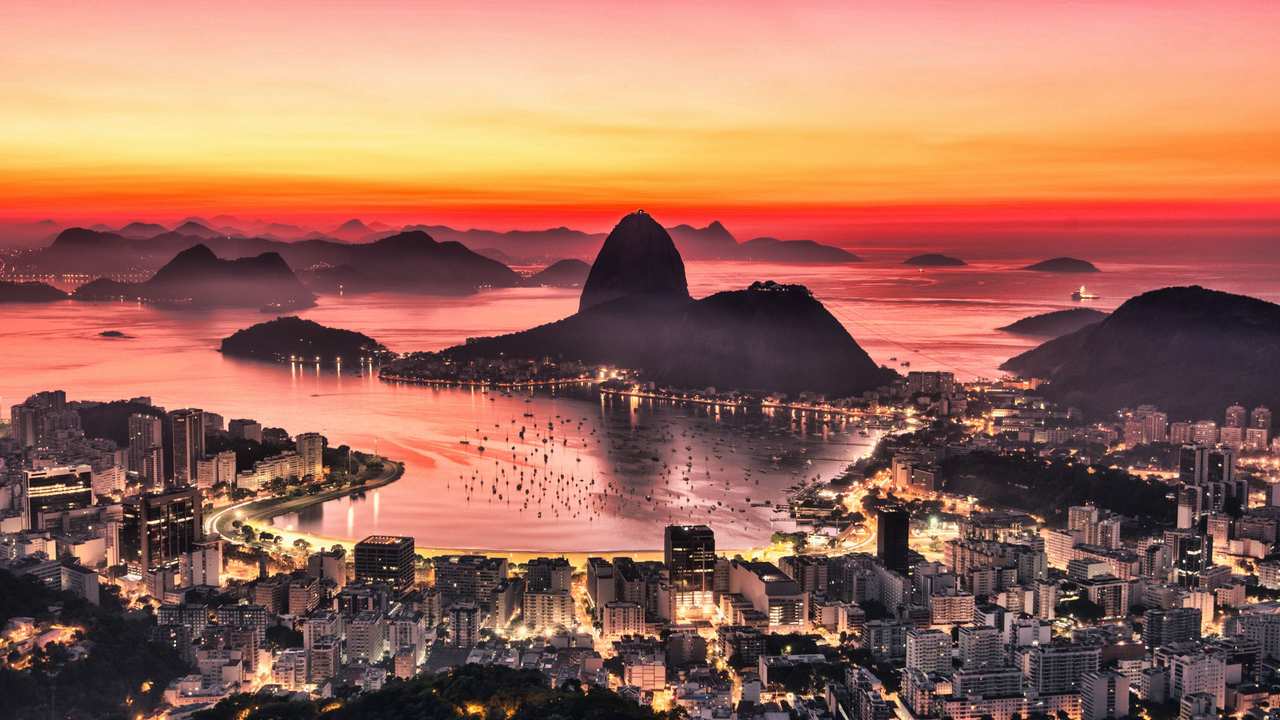 Vista panorâmica do Rio de Janeiro ao pôr do sol, com montanhas e cidade iluminada.