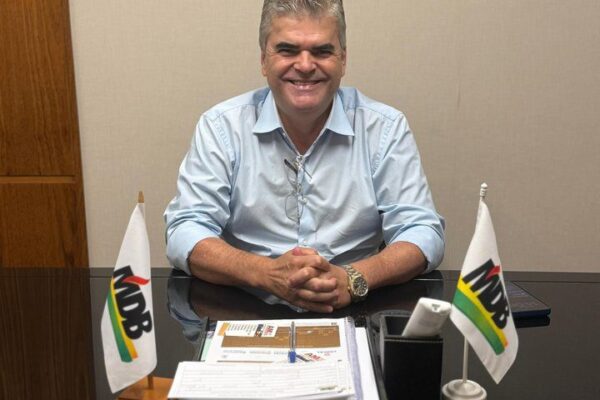 Washington Reis sorrindo em seu escritório, com bandeiras do MDB.