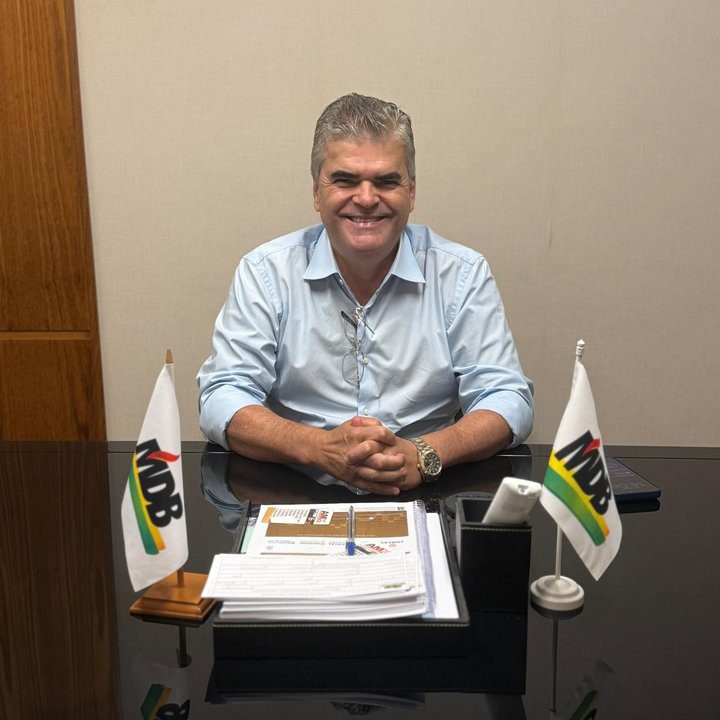 Washington Reis sorrindo em seu escritório, com bandeiras do MDB.