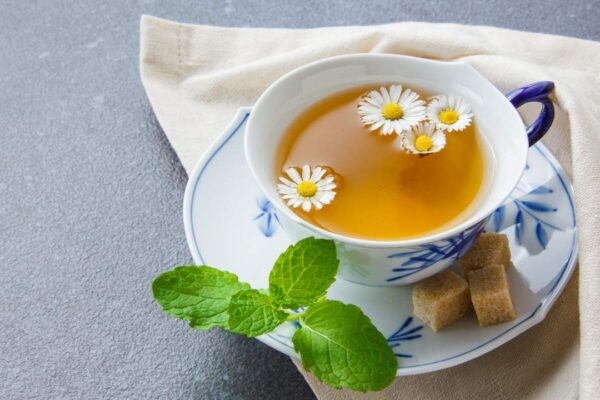 Xícara de chá de camomila com flores, acompanhada de açúcar e hortelã fresca.