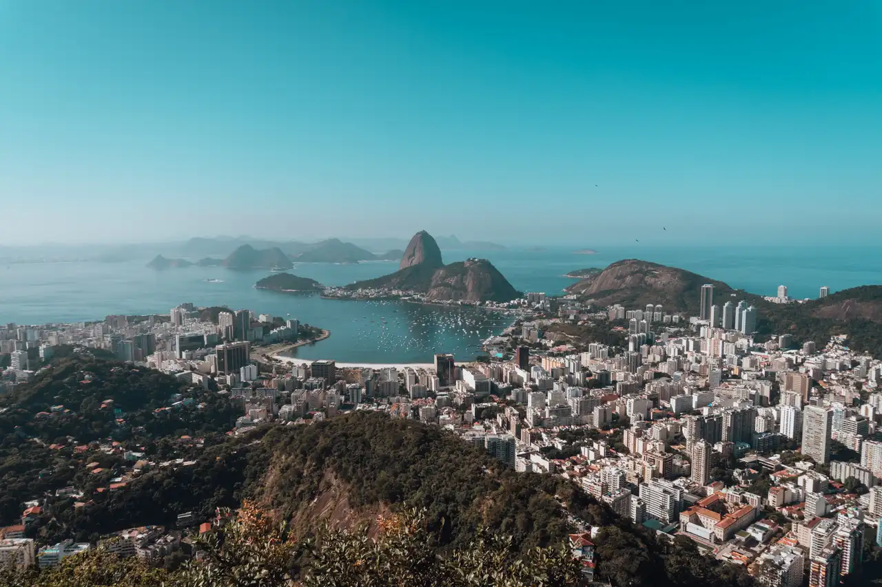 Imagem panorâmica do Rio de Janeiro, com prédios, montanhas, Baía de Guanabara e o Pão de Açúcar ao fundo em dia de céu claro.