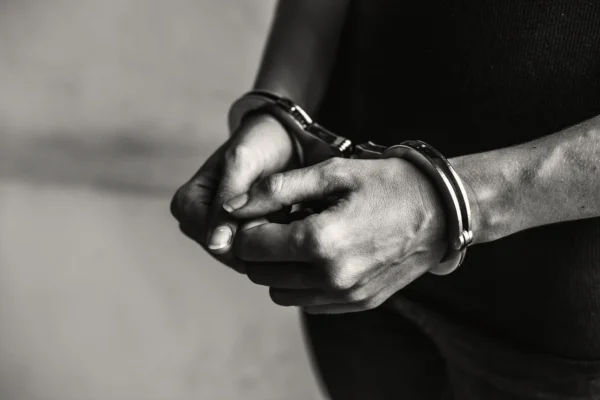 Homem aparece de costas, com as mãos algemadas, enquanto é acompanhado por um agente em uma rua de paralelepípedos.